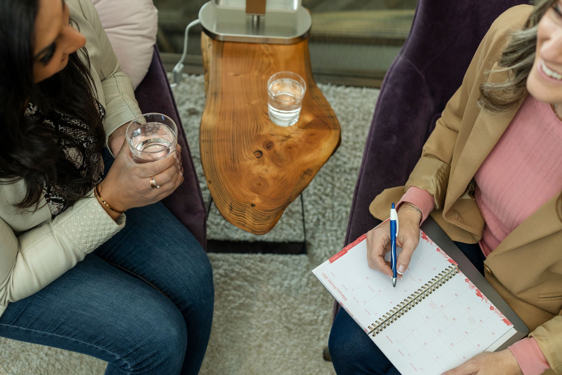 Two women are sitting in chairs talking and one is writing in a notebook.