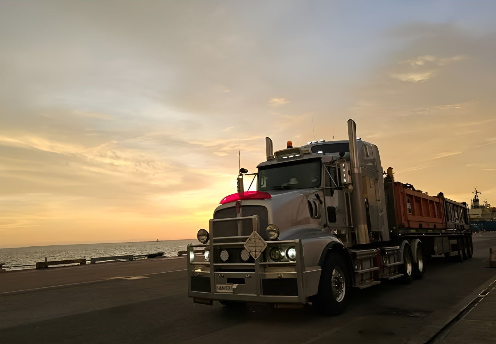 A Semi Truck is Driving Down a Road Near the Ocean at Sunset — Harker Haulage In East Arm, NT A Semi Truck is Driving Down a Road Near the Ocean at Sunset — Harker Haulage In East Arm, NT