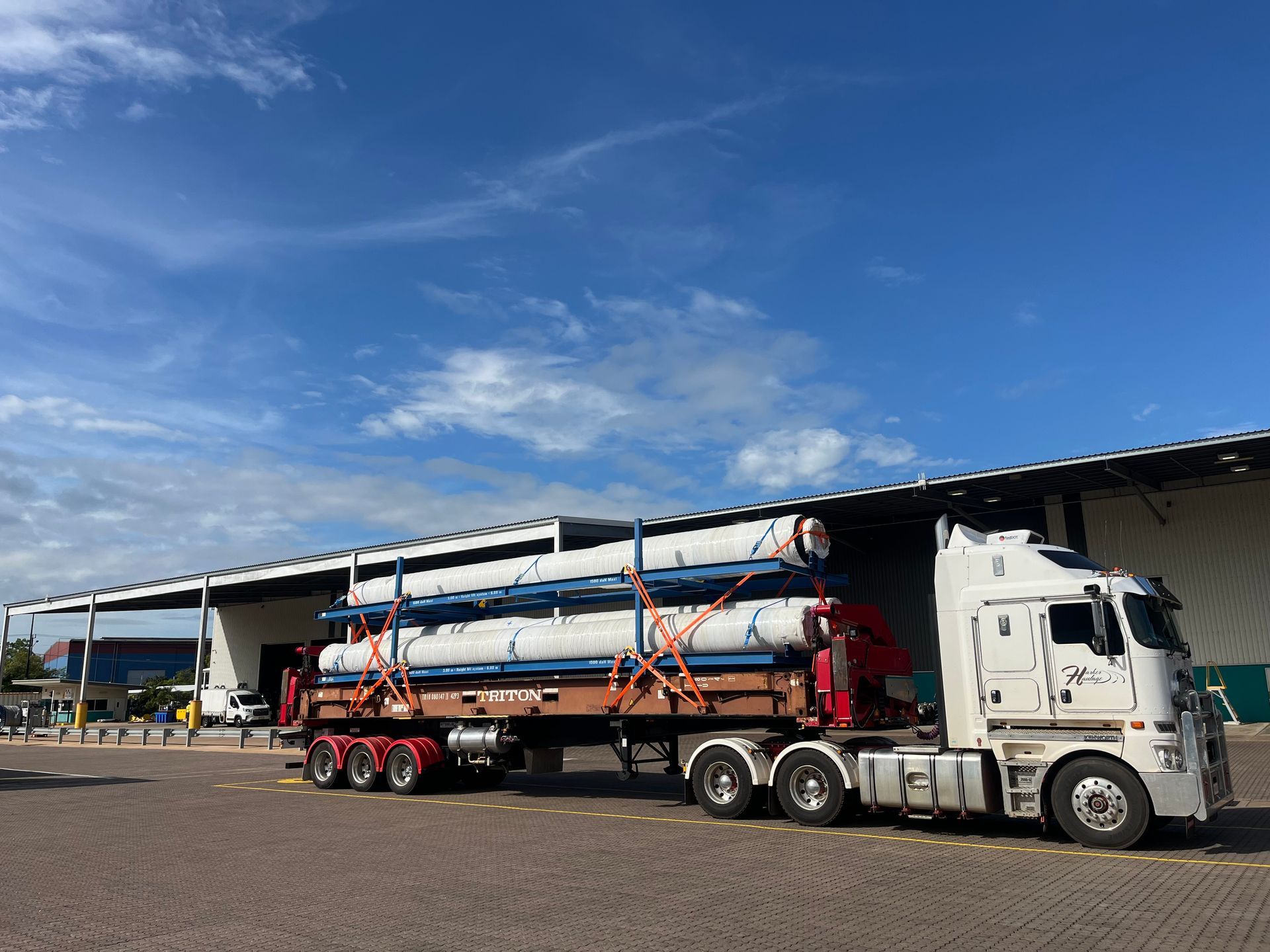 A semi-truck carrying large, white pipes, parked in front of a warehouse under a blue sky. — Harker Haulage In East Arm, NT