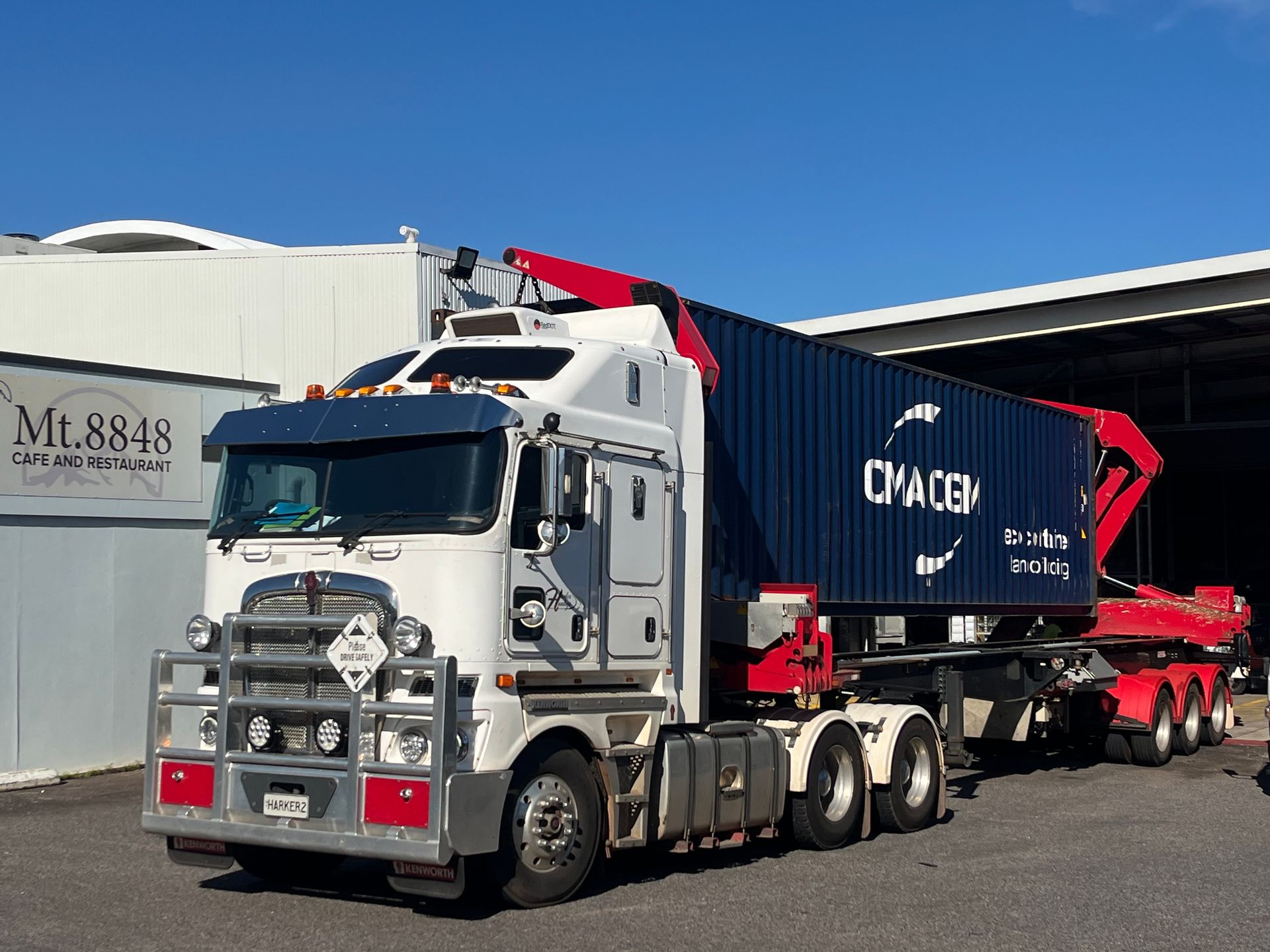White semi-truck with a blue cargo container; red crane arm, parked outside a building under a blue sky. — Harker Haulage In East Arm, NT