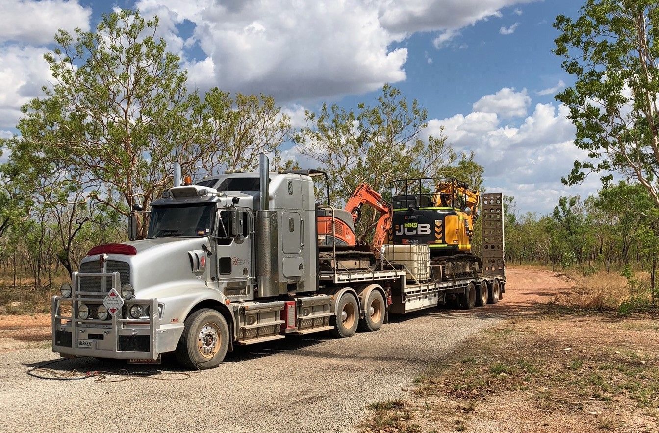 Silver semi-truck hauling construction equipment on a dirt road, trees in the background under a blue sky. — Harker Haulage In East Arm, NT
