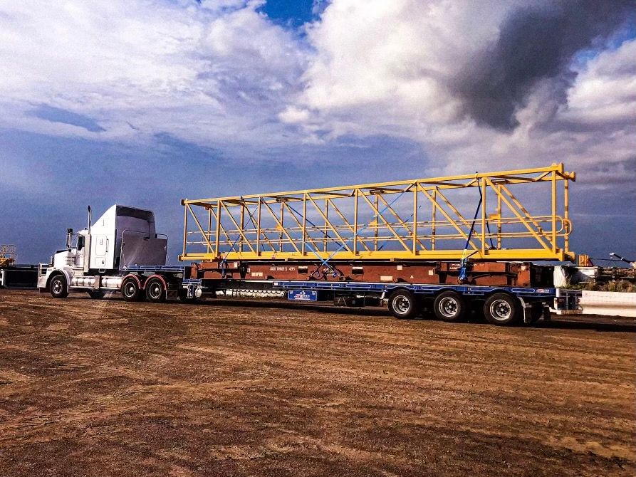White semi-truck hauling a long, yellow metal structure on a blue trailer across a dirt lot under a cloudy sky. — Harker Haulage In East Arm, NT