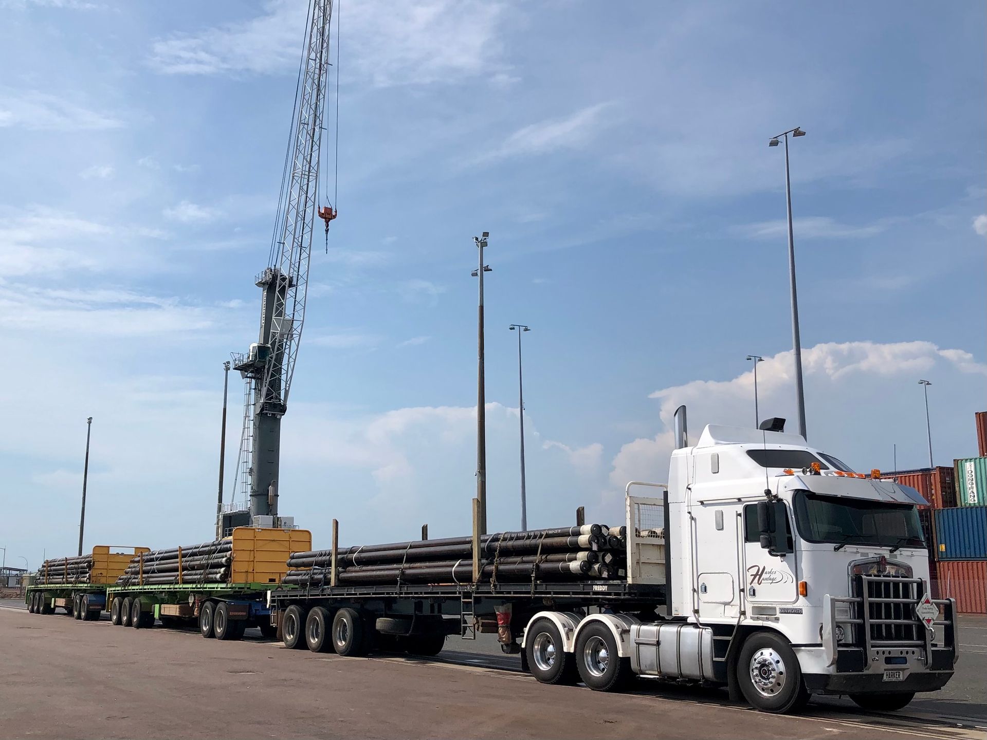 White semi-truck hauling long pipes at a port with a crane and blue sky. — Harker Haulage In East Arm, NT