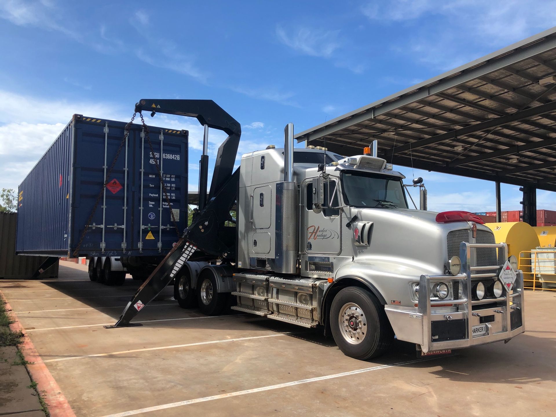 Silver semi-truck lifting a blue shipping container. Outdoors on a sunny day. — Harker Haulage In East Arm, NT