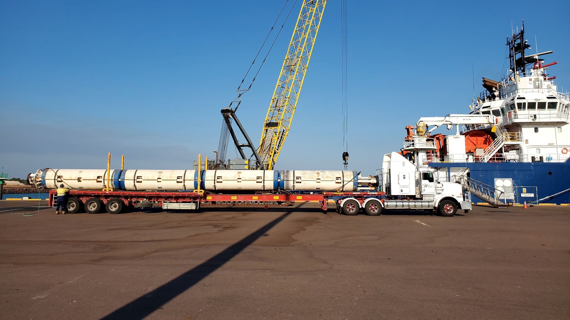 A semi-truck hauls cylindrical cargo at a dock, with a crane and blue ship in the background under a sunny sky. — Harker Haulage In East Arm, NT