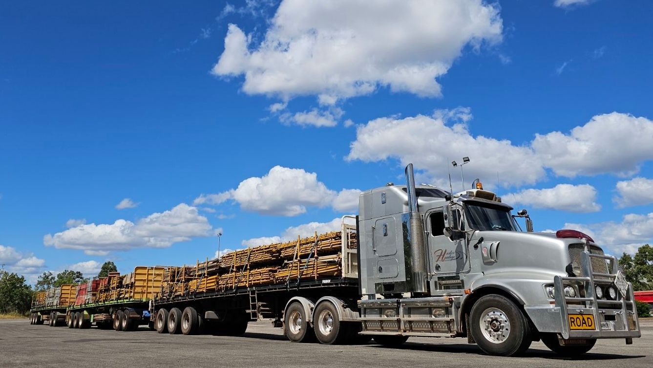 A large truck hauling logs on a flatbed trailer under a blue sky with puffy clouds. — Harker Haulage In East Arm, NT
