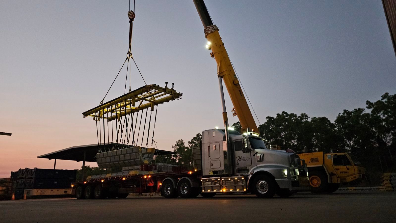 A crane lifts metal equipment from a flatbed truck in a dusk setting, with a white semi-truck. — Harker Haulage In East Arm, NT