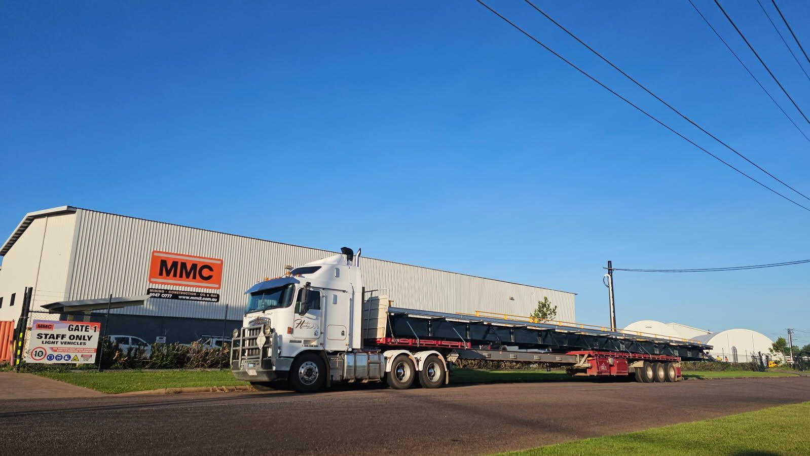 White semi-truck with flatbed trailer loaded with metal, parked in front of a warehouse under a blue sky. — Harker Haulage In East Arm, NT
