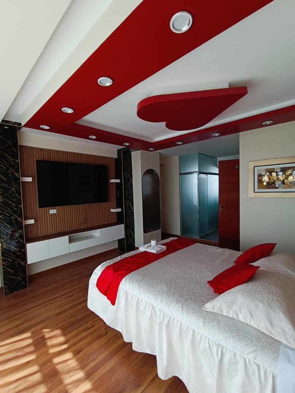 Dormitorio con decoración en rojo y blanco, piso de madera, cama, TV y acento de techo en forma de corazón.