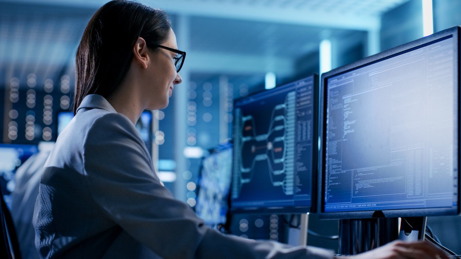 A woman is sitting in front of two computer monitors in a server room.