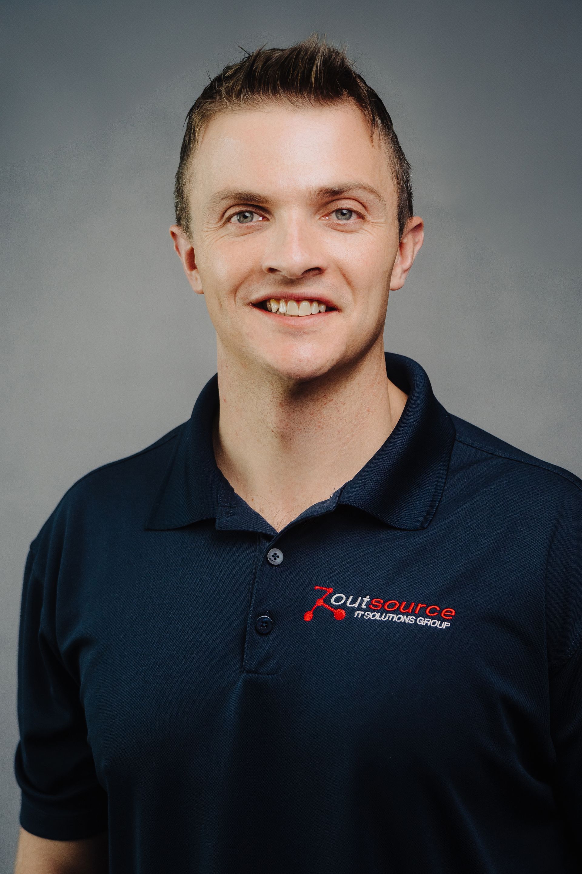 Man wearing a navy polo shirt with logo, smiling. Gray background.