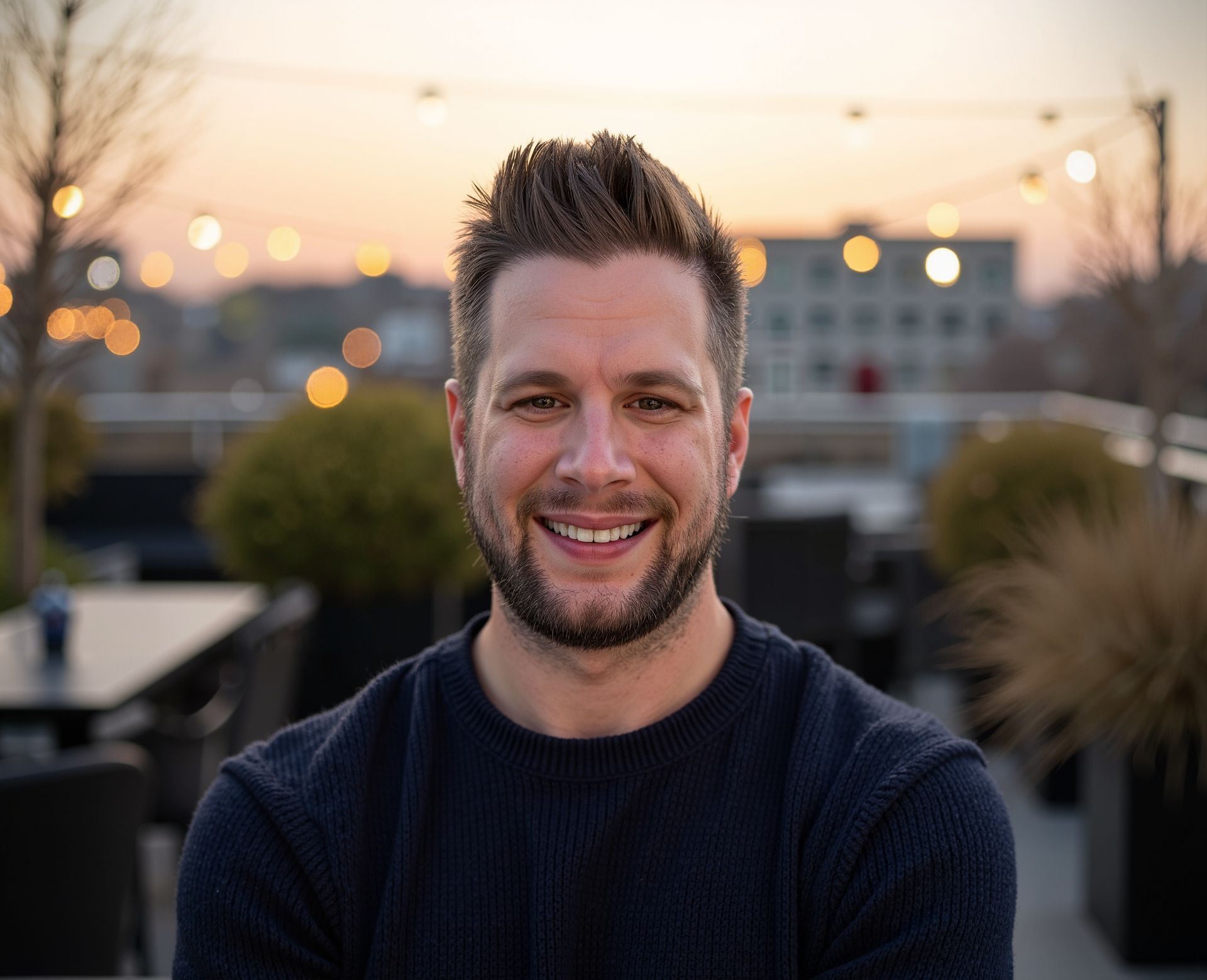 Man with short brown hair smiling, outdoors at dusk. He's wearing a dark sweater, with blurred lights and buildings in background.