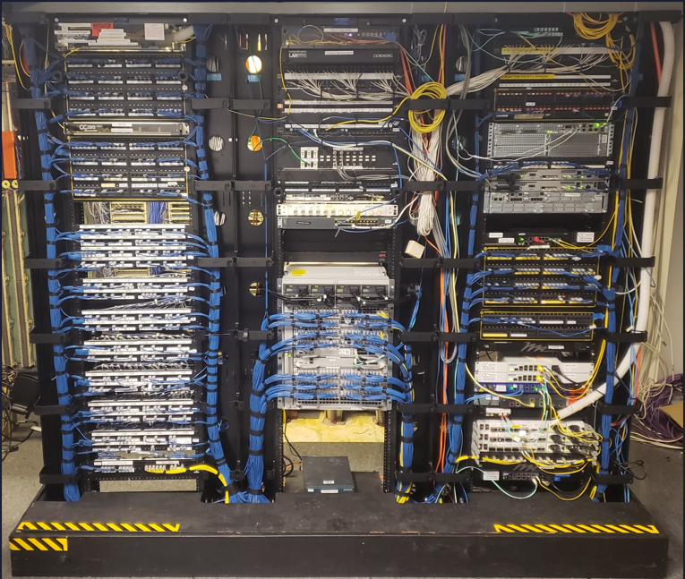 Server racks filled with network equipment, cables, and patch panels, with yellow and black hazard stripes at the base.
