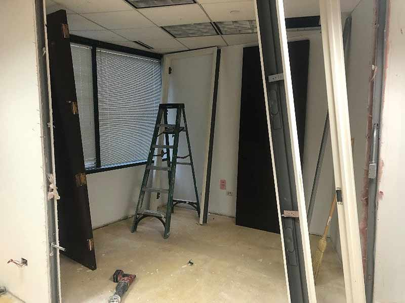 Office room under renovation; walls open, ladder present, dark doors leaning against walls.