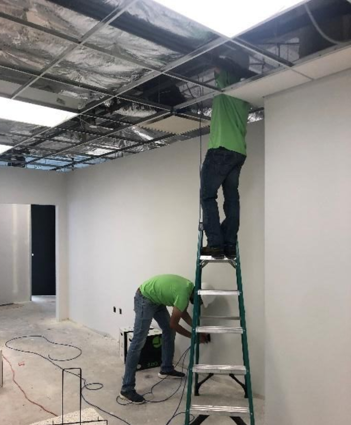 Two men in green shirts work on a ceiling, one on a ladder, in a room under construction.
