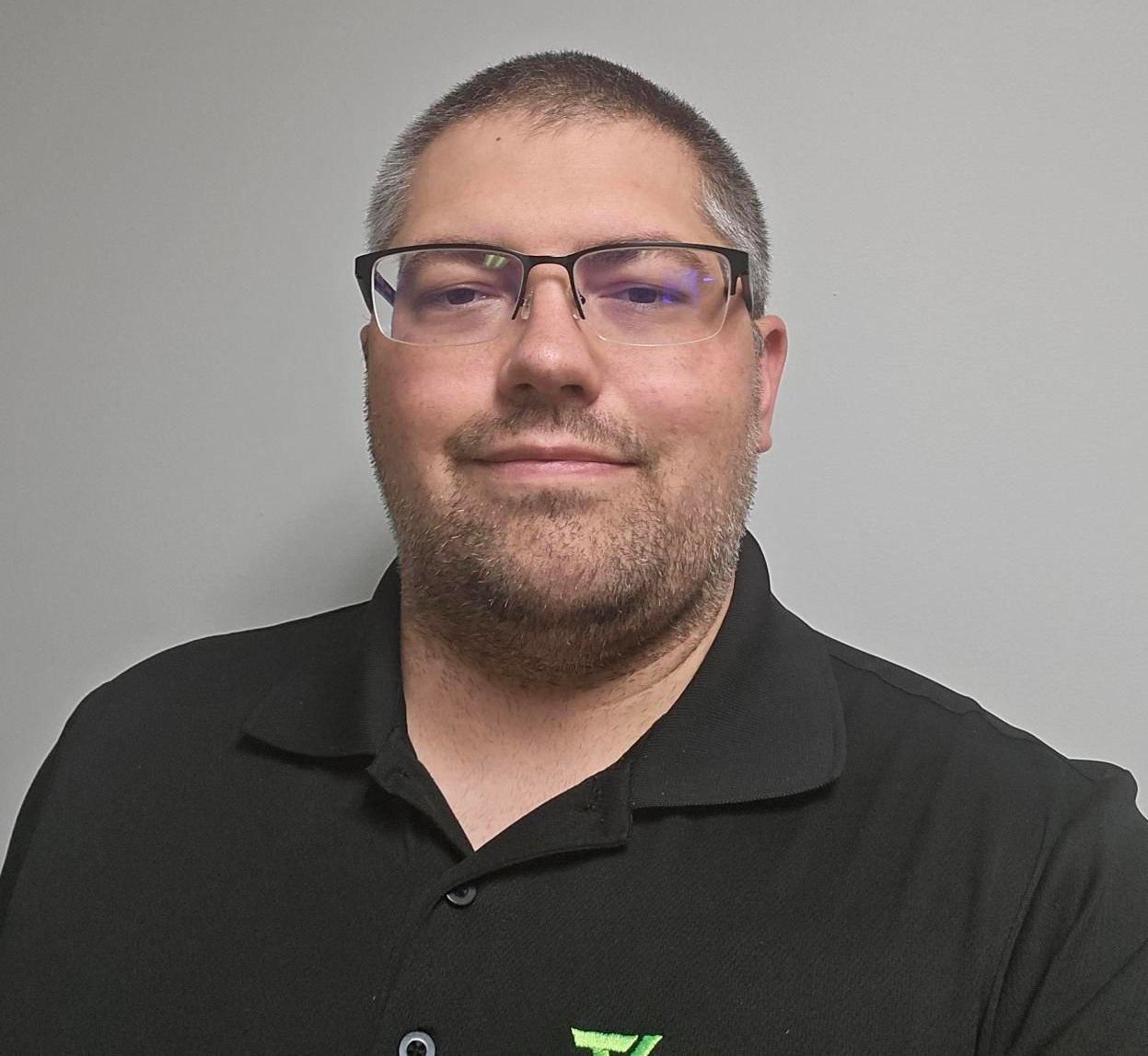 Man with glasses and black polo shirt smiles in front of a gray wall.