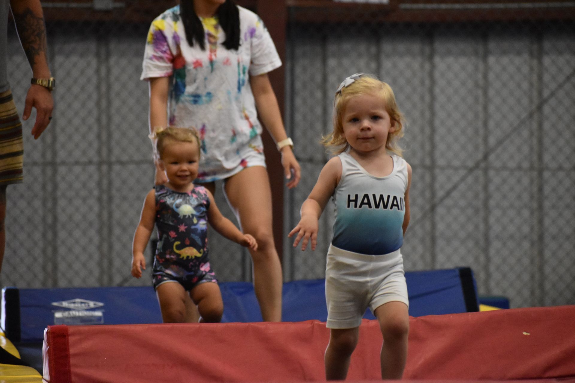 Two toddlers in leotards walking near a woman in a gym. One has 