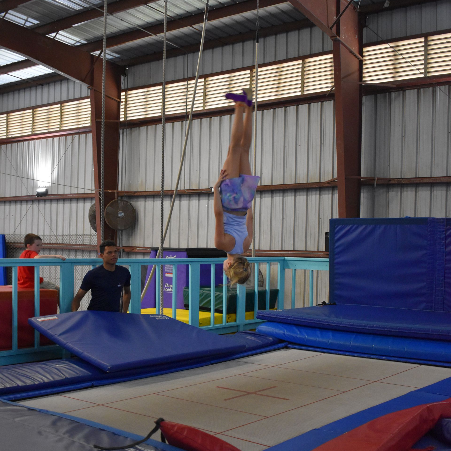 Girl in blue and purple clothing upside down on a trampoline with instructor spotting with a mat.
