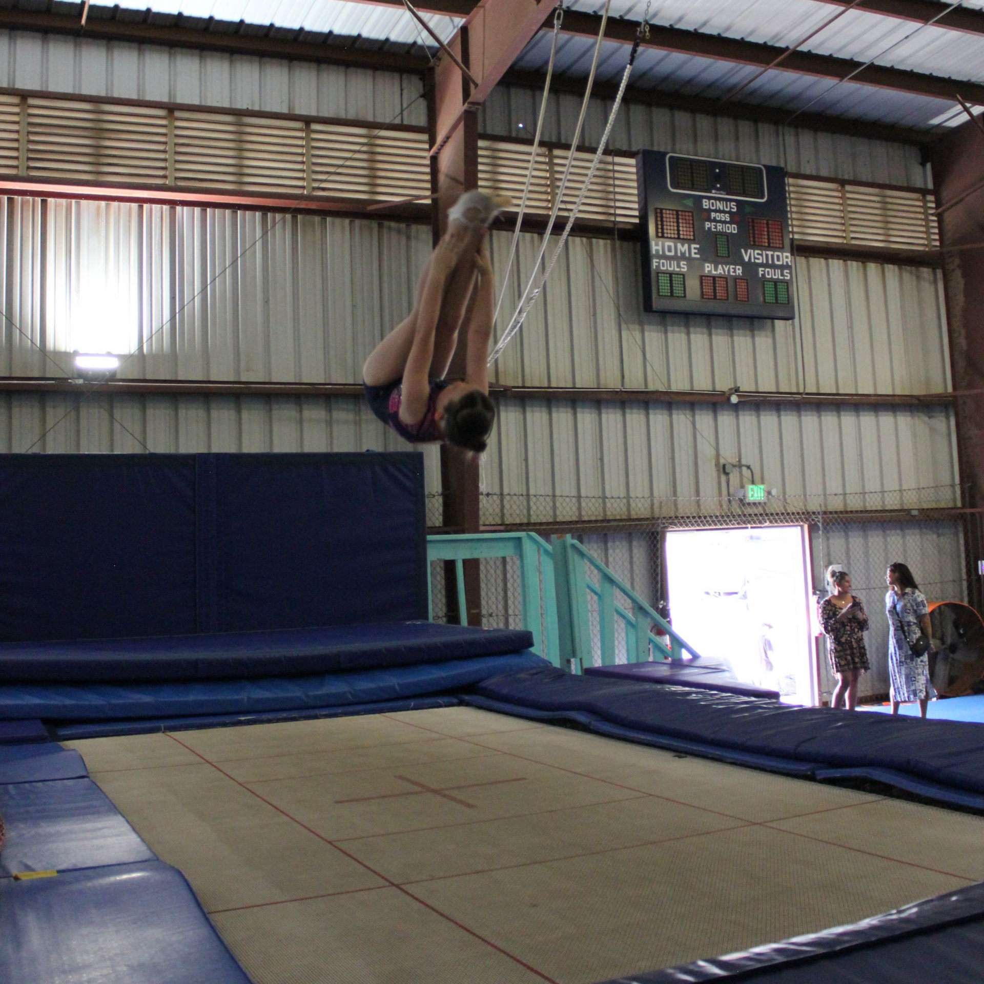 Girl in purple leotard jumps on trampoline in a gym. Other gymnasts, blue mats, American flag.