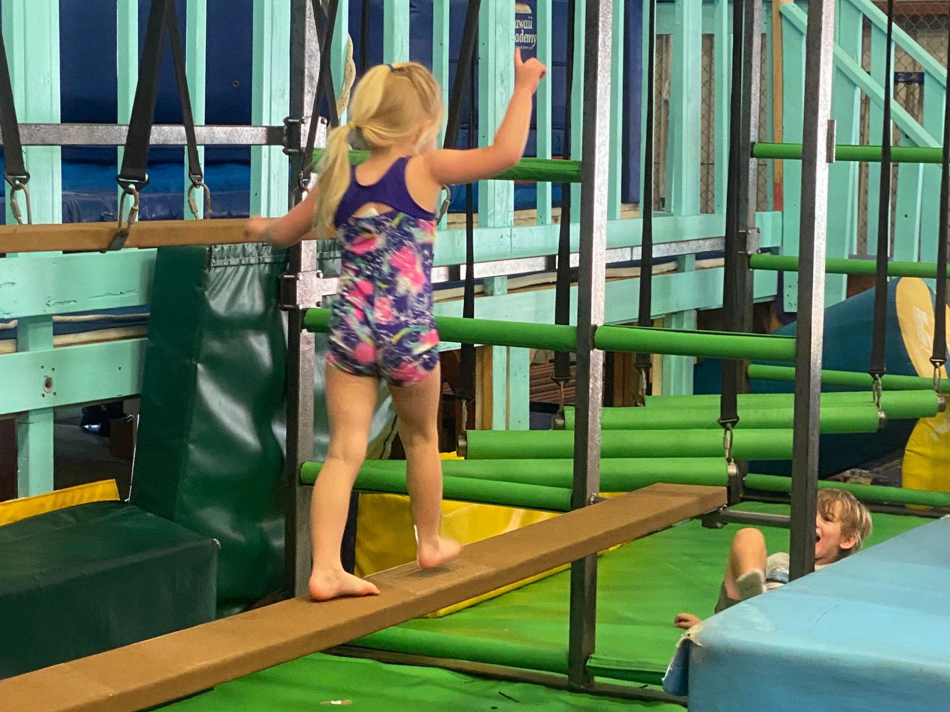 Blonde girl walks across a balance beam in an indoor gym with other children playing.