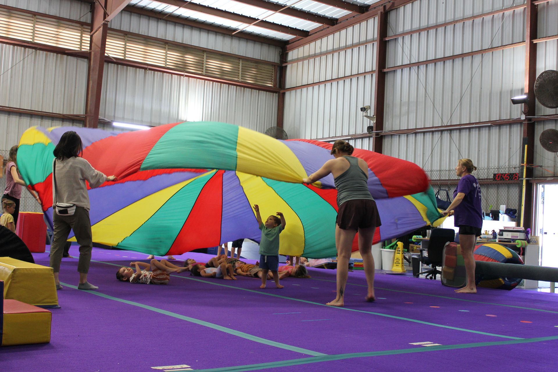Two toddlers in leotards walking near a woman in a gym. One has 