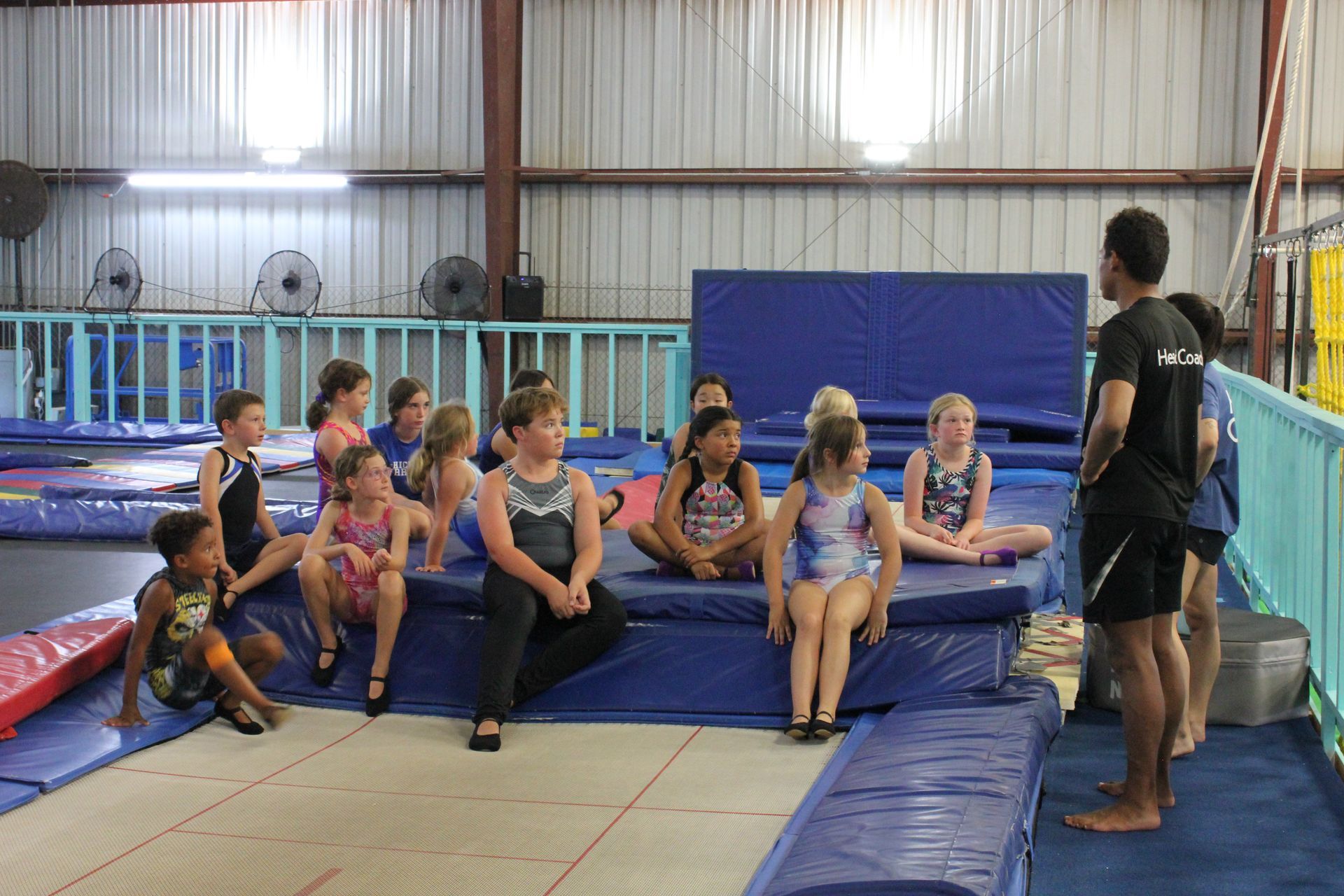 Group of children on blue mats, listening to a coach in a gym with trampolines.