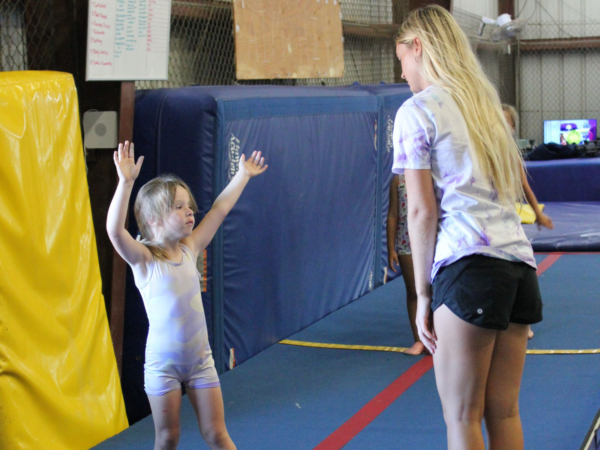 Young girl practicing gymnastics on a blue mat, arms raised. Coach watches. Indoor gym setting.