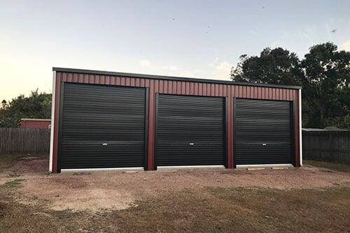 A Garage With Three Black Garage Doors Is Sitting On Top Of A Dirt Field — Dunnrite Mareeba Sheds & Garages In Mareeba, QLD