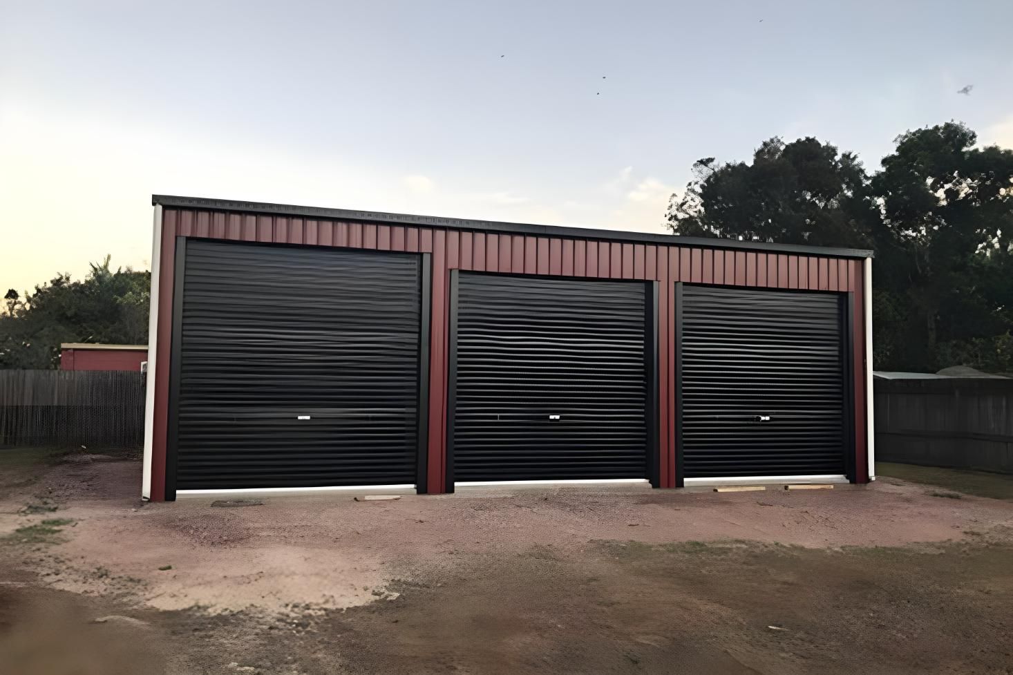A Red Garage With Three Black Garage Doors In A Dirt Lot — Dunnrite Mareeba Sheds & Garages In Mareeba, QLD