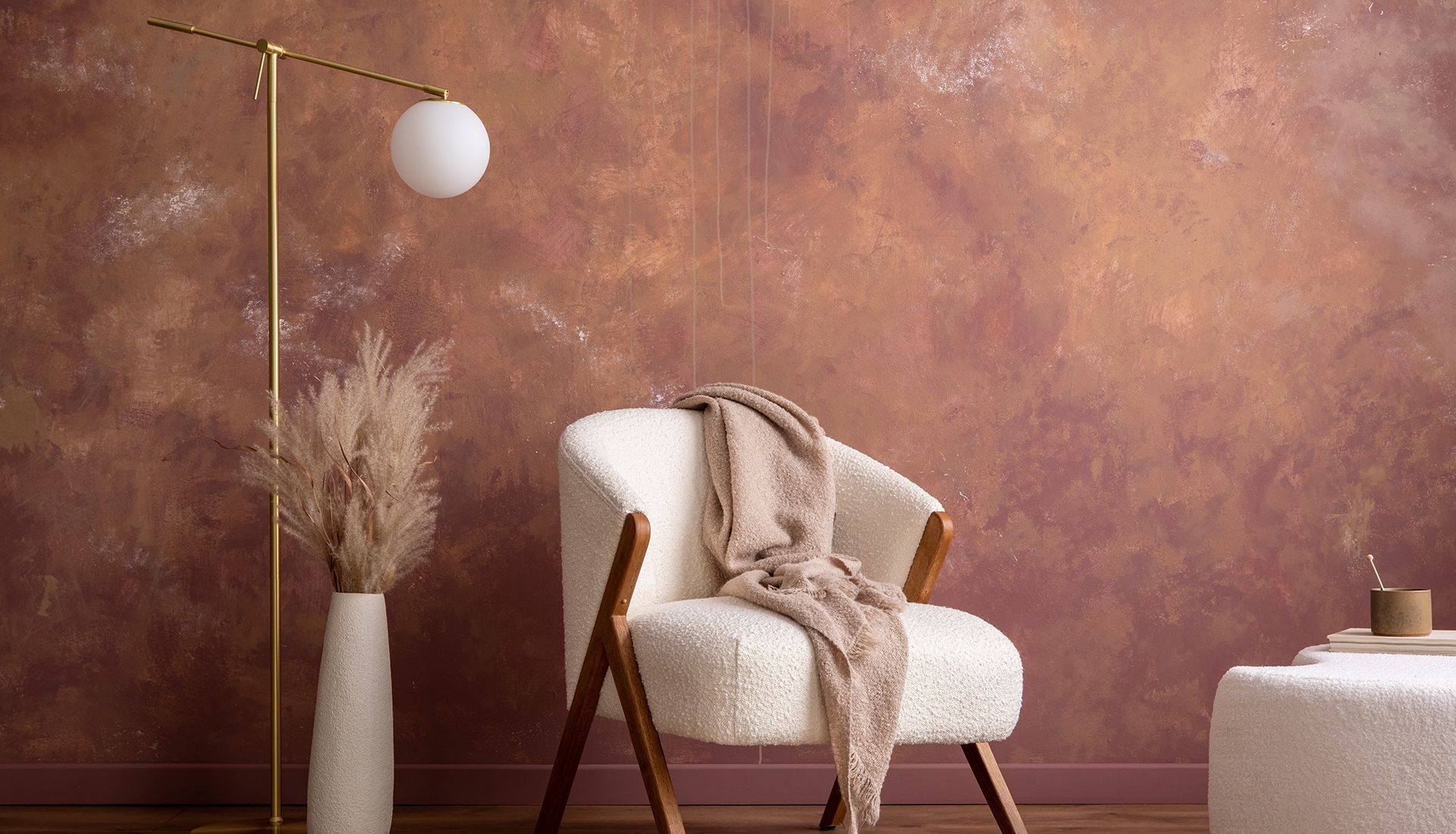 Cozy room with textured red-brown wall, cream chair, tan blanket, gold lamp, vase with dried plants.