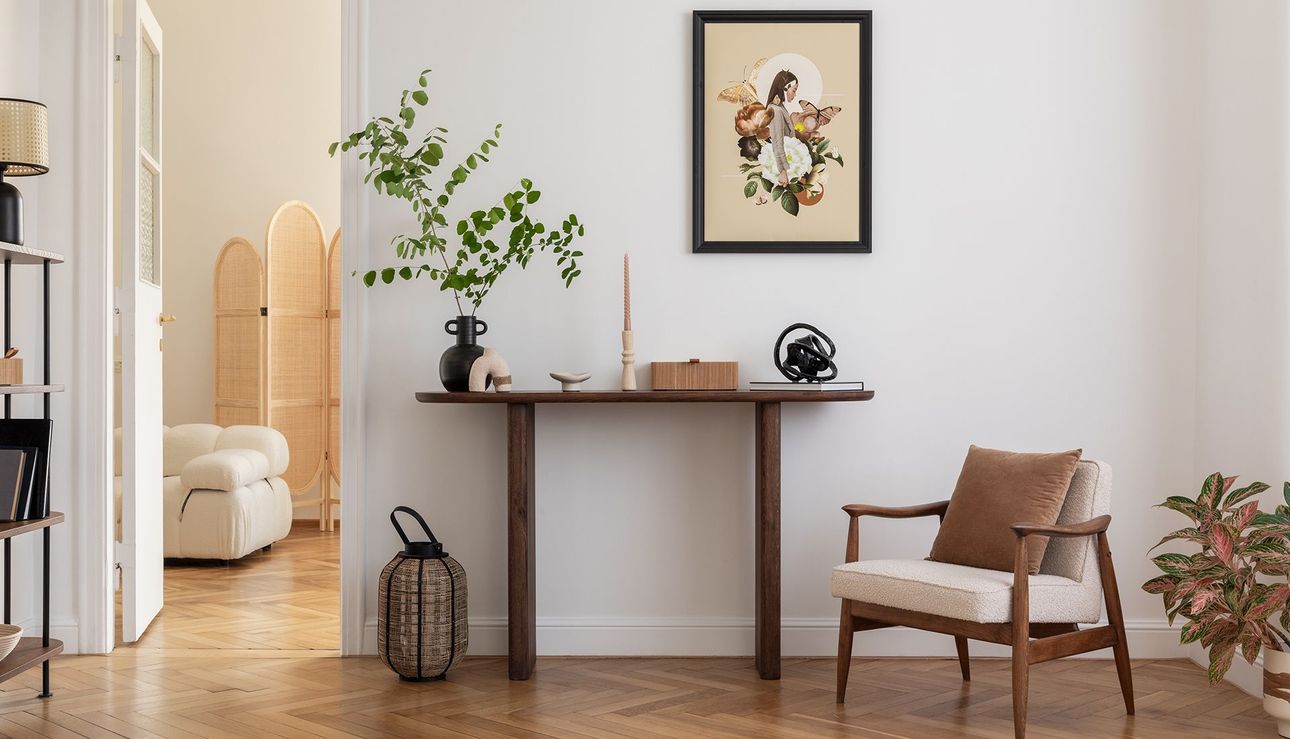 Cozy living space with a dark wood console table, artwork, chair, and greenery against a white wall.