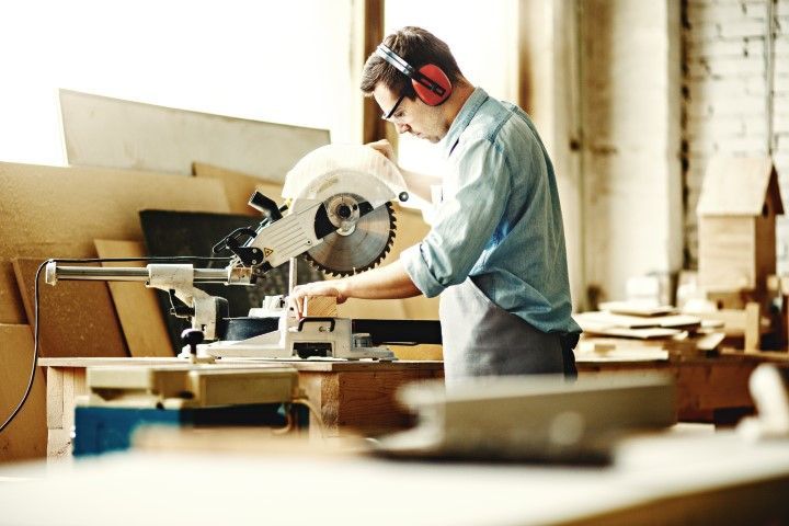 Man operating a miter saw in a workshop, wearing earmuffs and safety glasses, cutting wood.