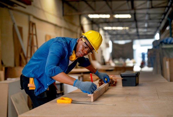 Carpenter in a workshop wearing a hard hat, measuring wood with a pencil.
