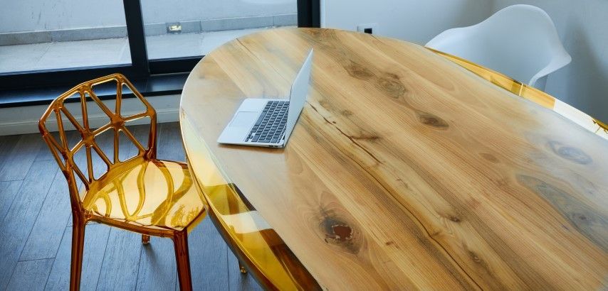 A wooden oval table with a laptop and two chairs, one orange and one white, near a window.