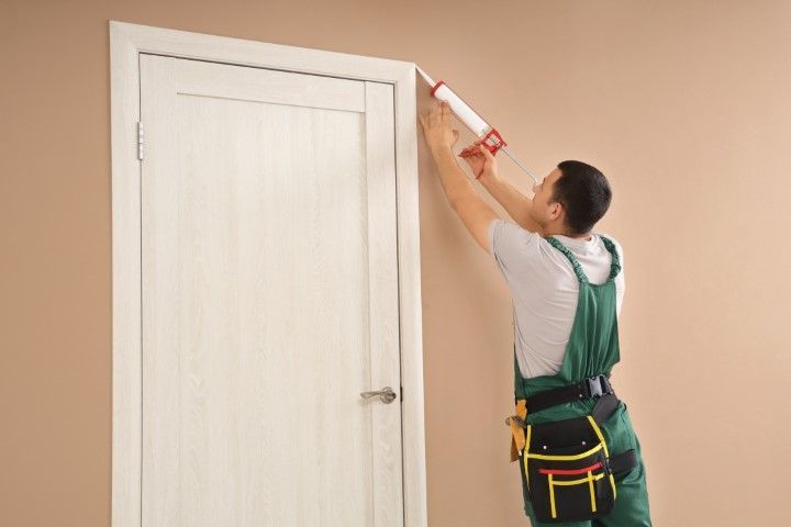 Person in green overalls caulking around a white door frame on a tan wall.