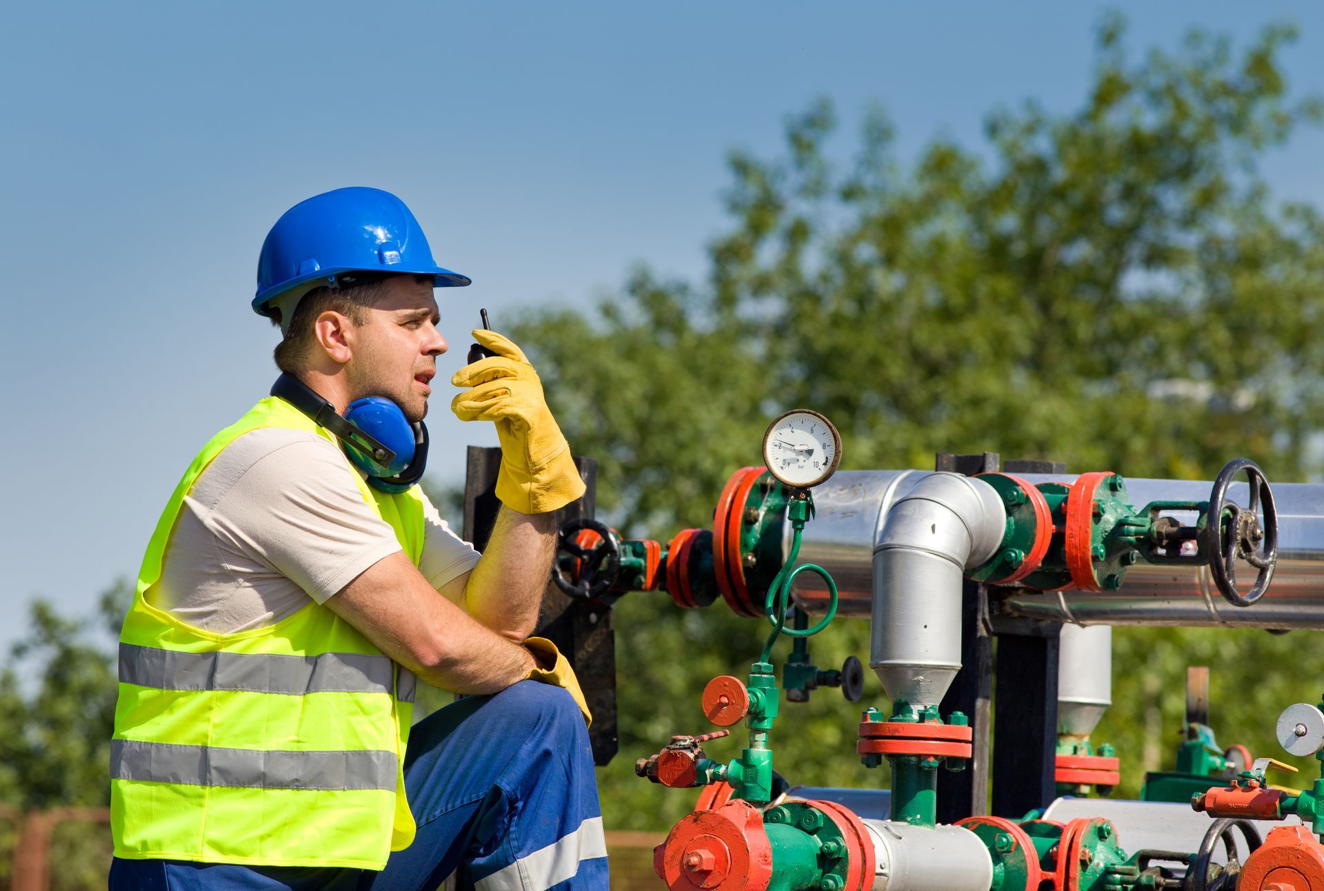 Man in safety gear operating a radio next to industrial pipes outdoors under a blue sky.