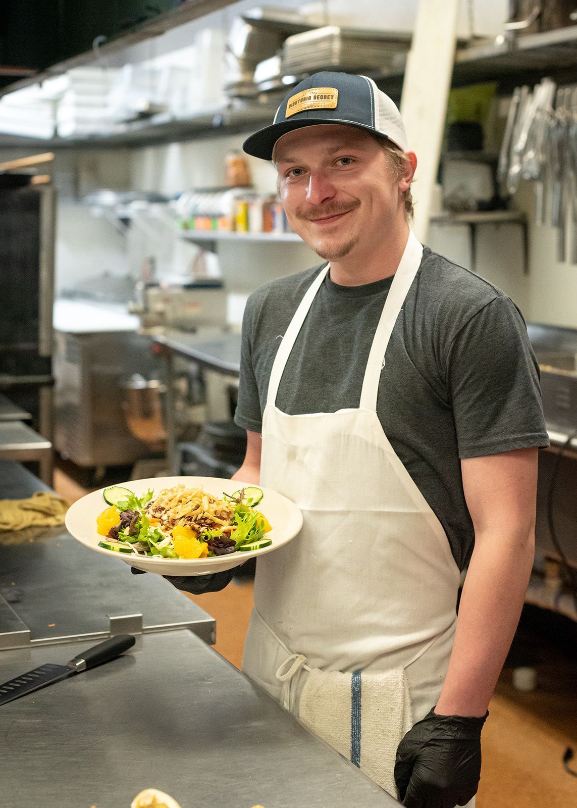 A man in an apron is holding a plate of food in a kitchen.