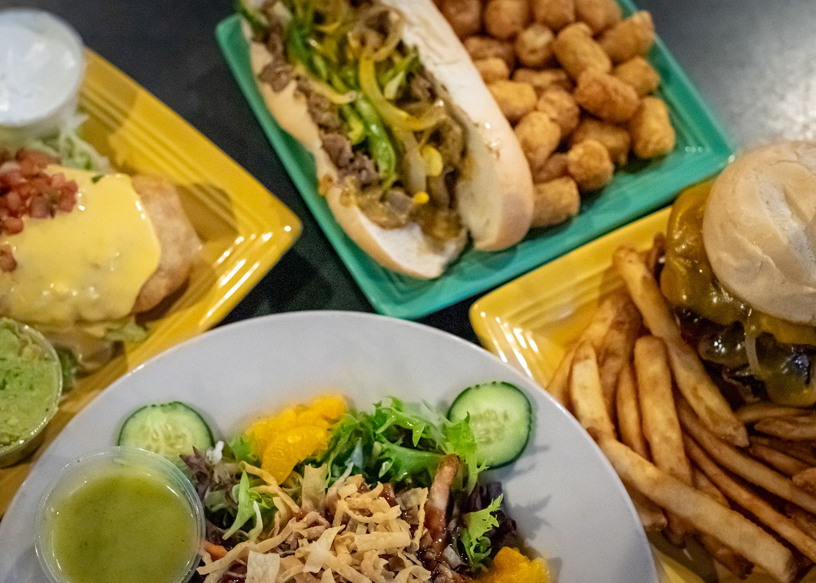 A table topped with plates of food including a hamburger , Philly, salad and french fries.
