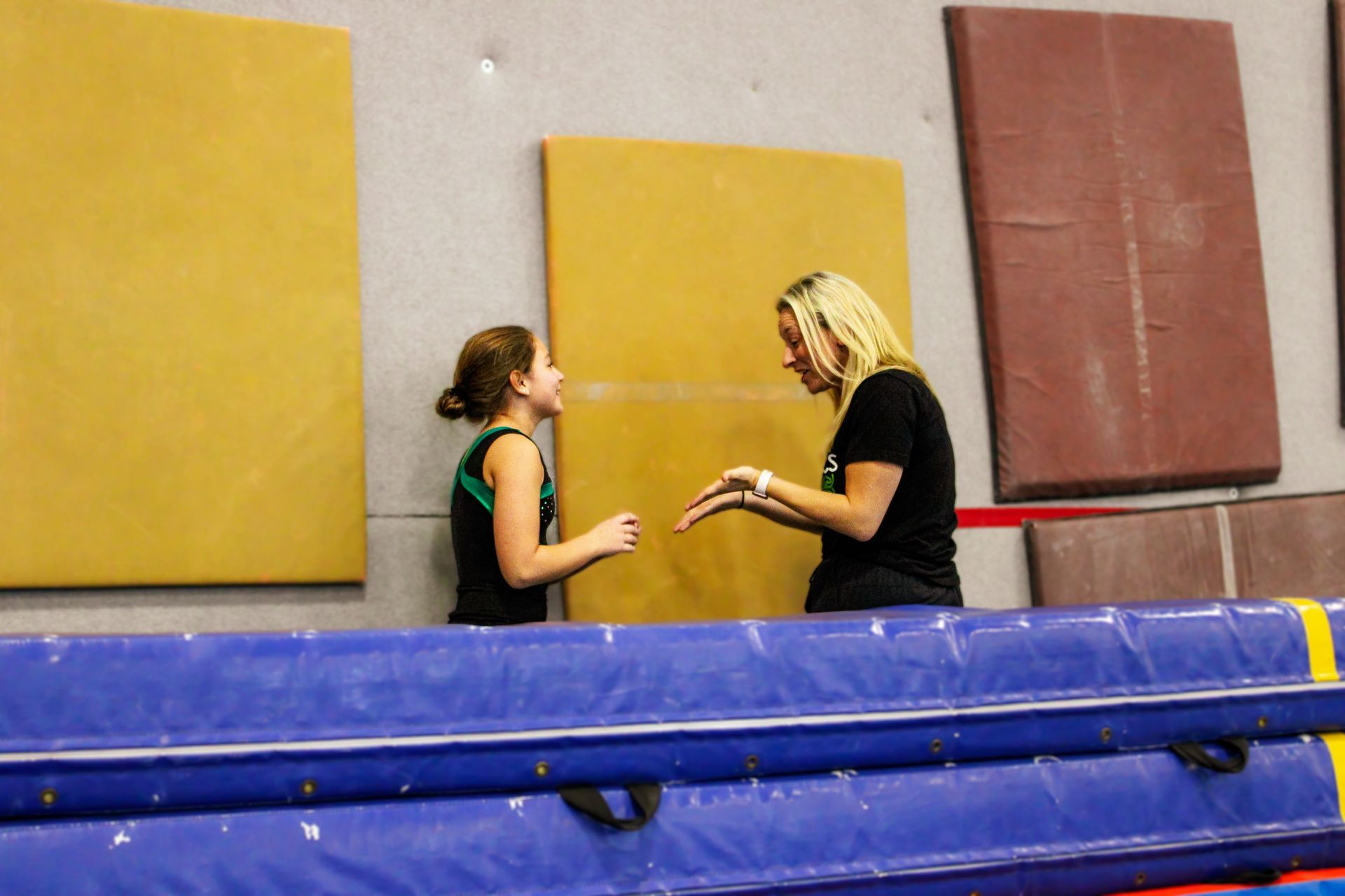 Two people talking beside a blue mat in a gym, with yellow and red wall pads behind them.