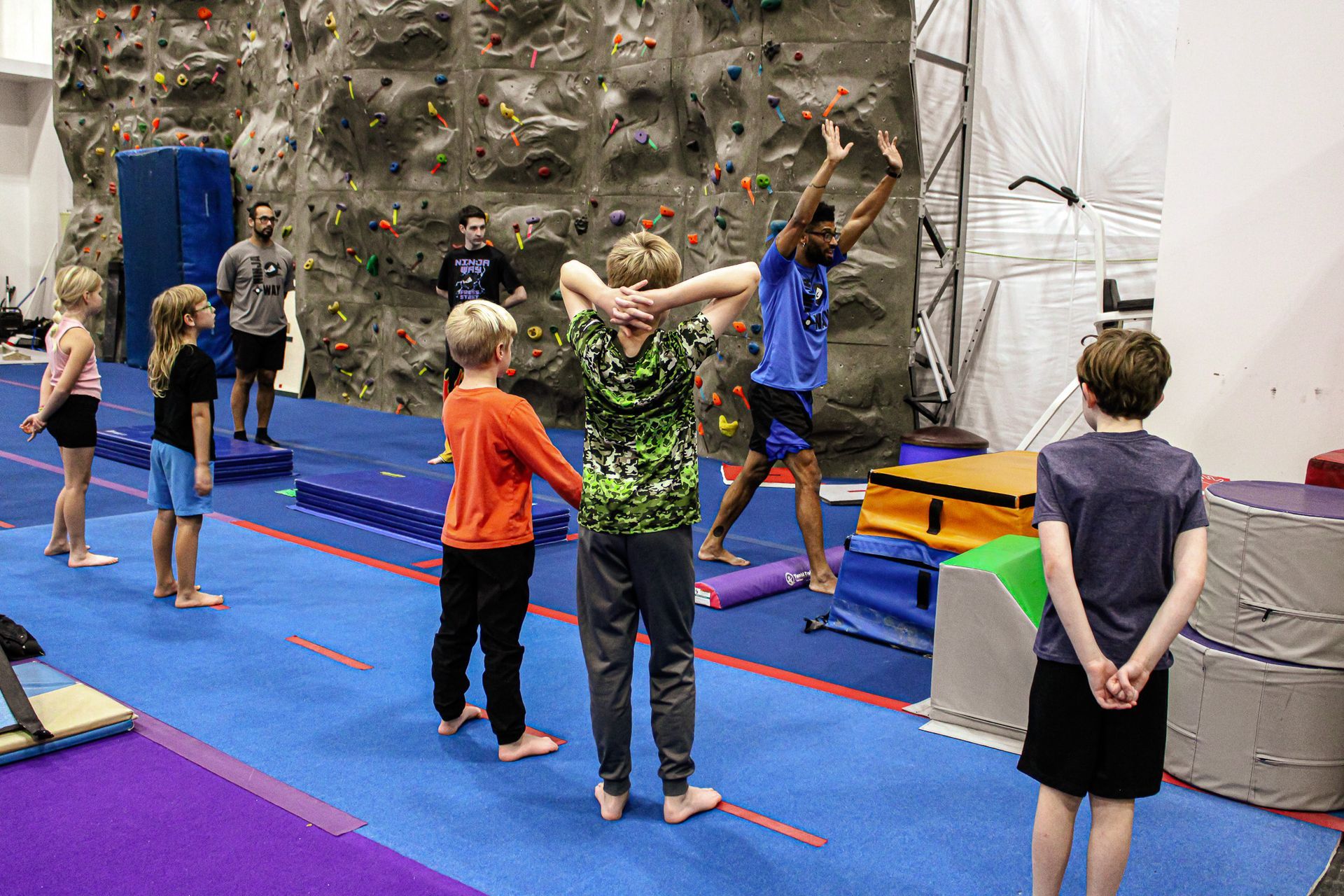 Children practicing gymnastics in a colorful indoor gym, stretching on blue mats with one instructor demonstrating a pose.