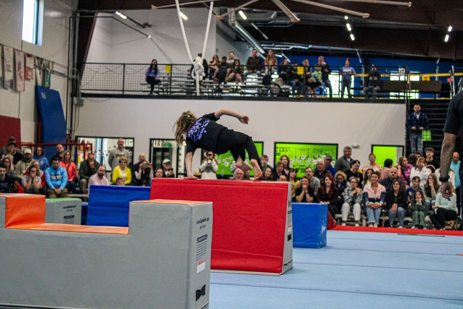 Gymnast balancing on a red foam block in a crowded indoor competition arena