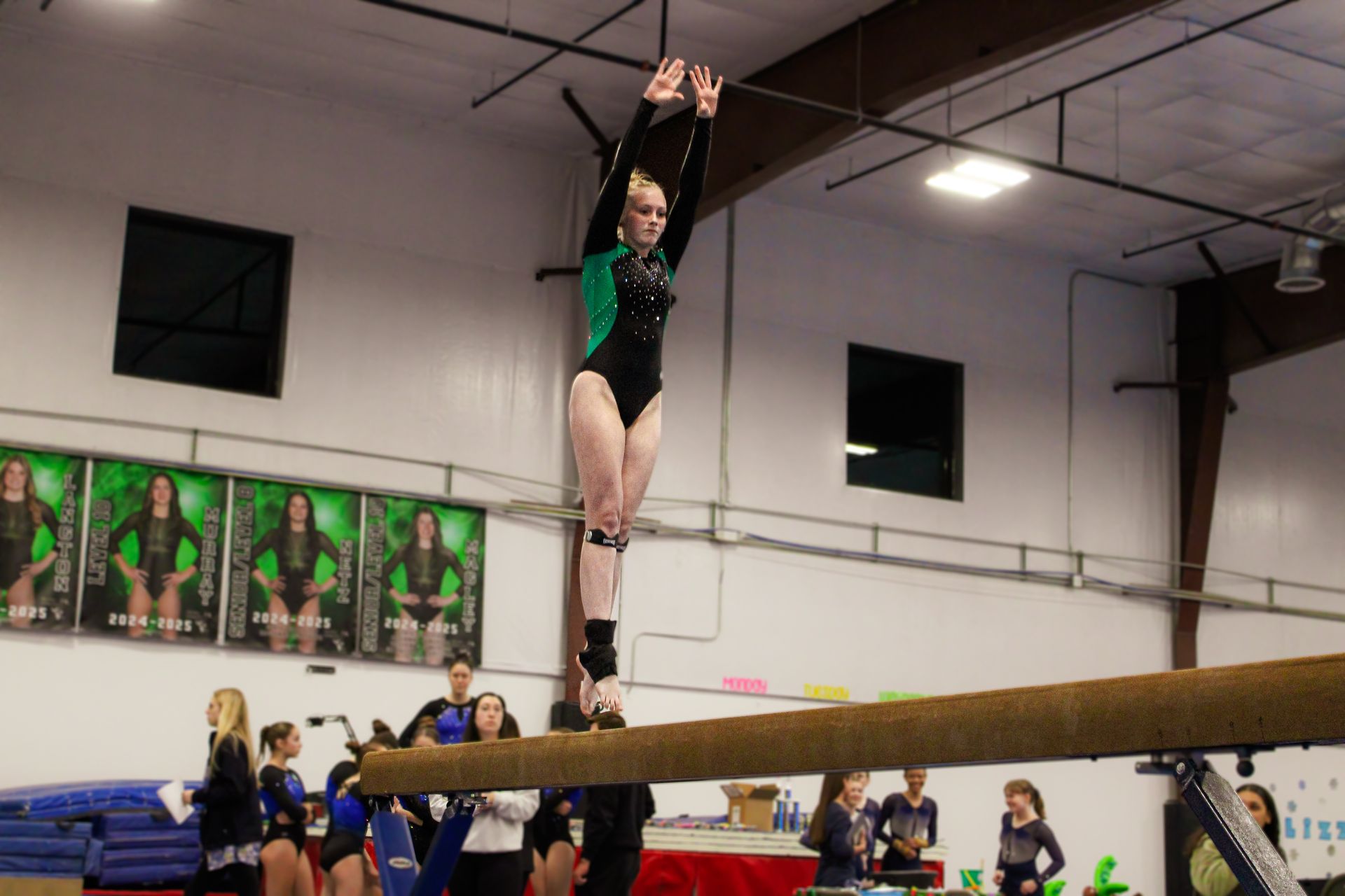 Gymnast balancing on a beam indoors, arms raised overhead in a black and green leotard.