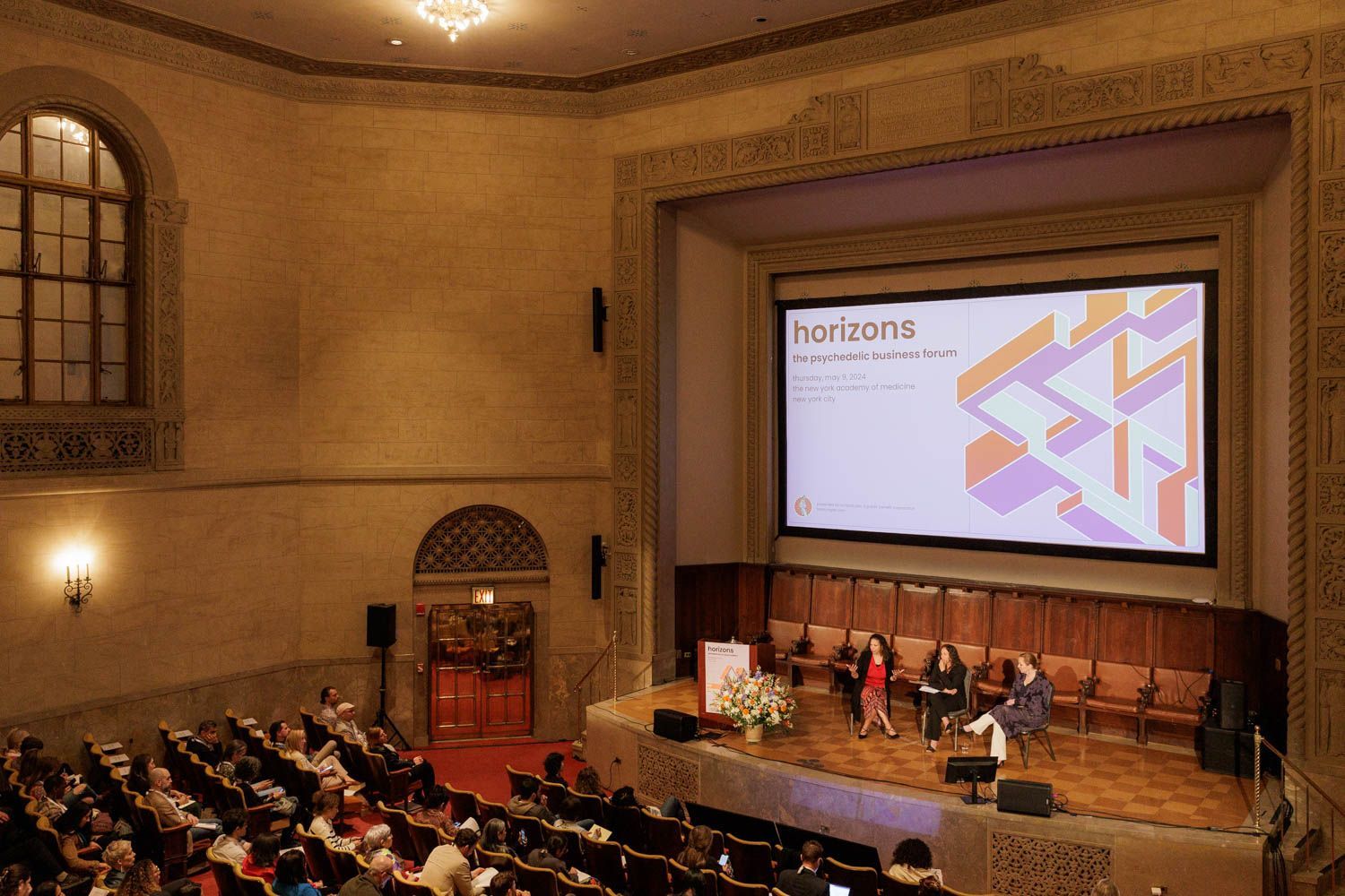 Audience watches panel discussion on a stage in a large auditorium; screen displays "horizons" graphic.