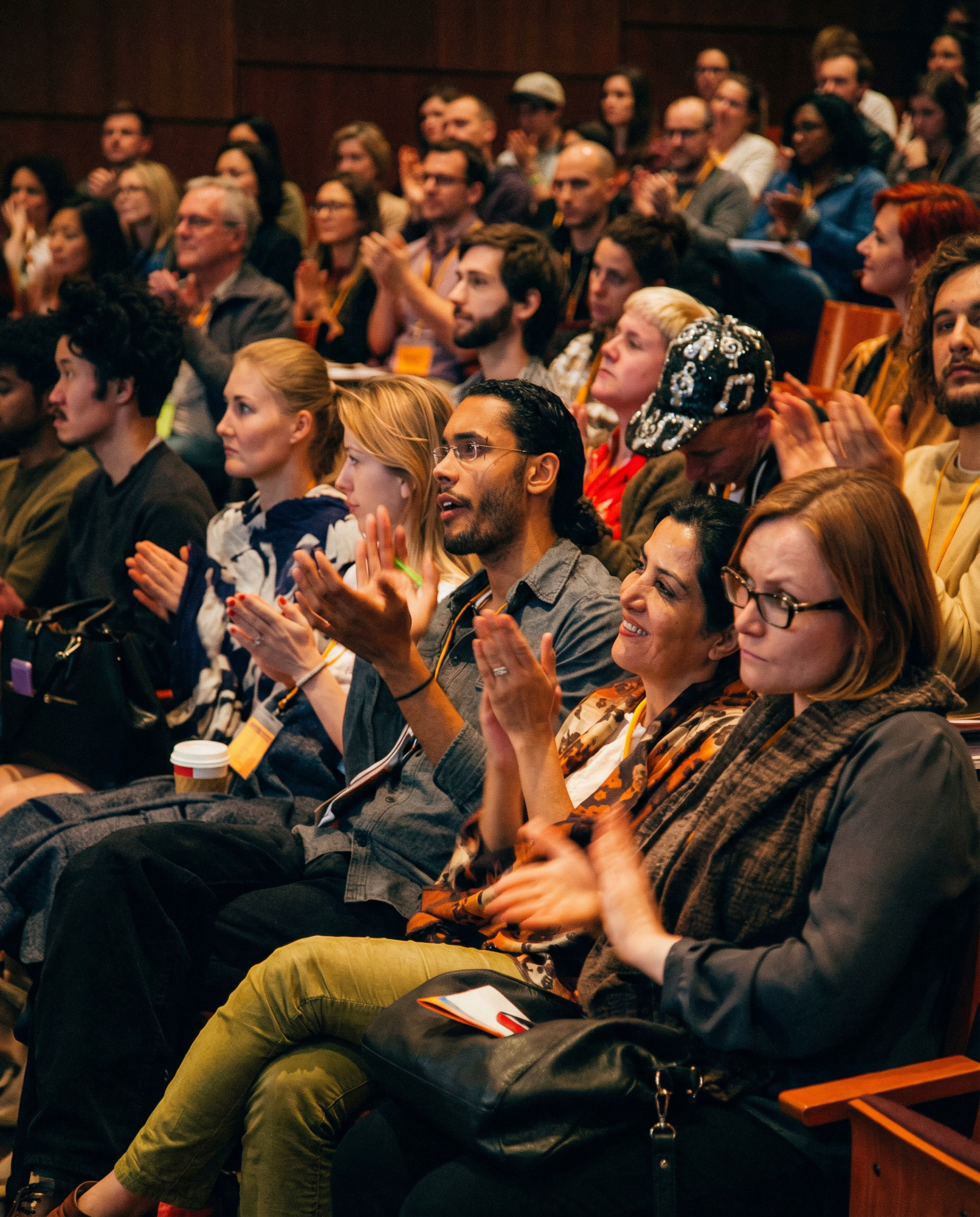 Audience clapping enthusiastically in a theater. People in various attire. Warm lighting.
