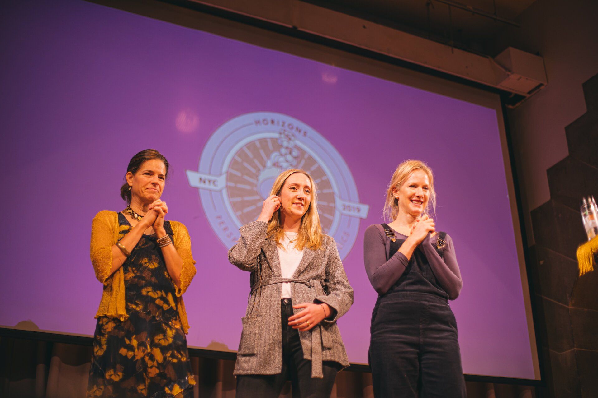 Three women are standing on a stage in front of a large screen.