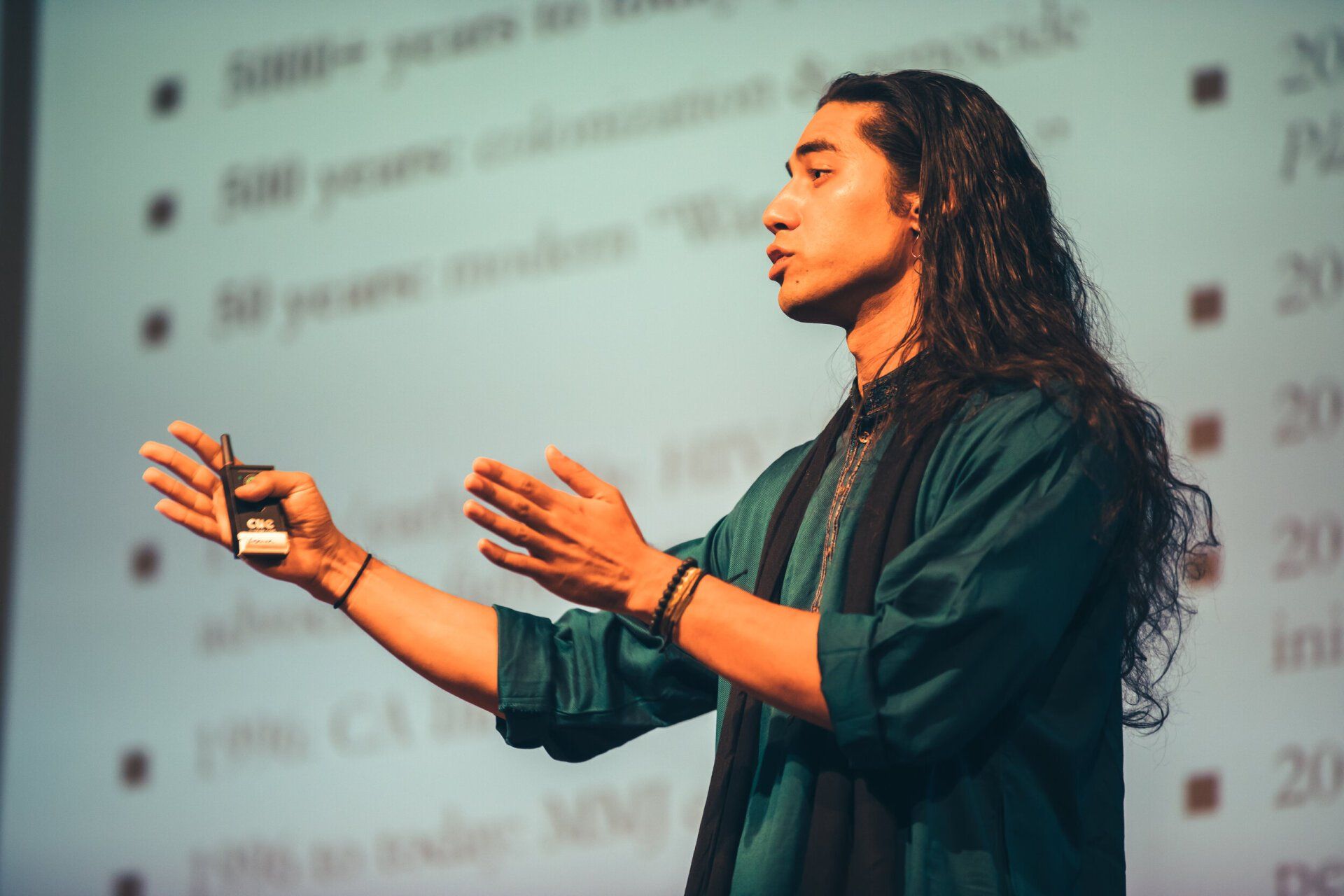 A man with long hair is giving a presentation in front of a screen.