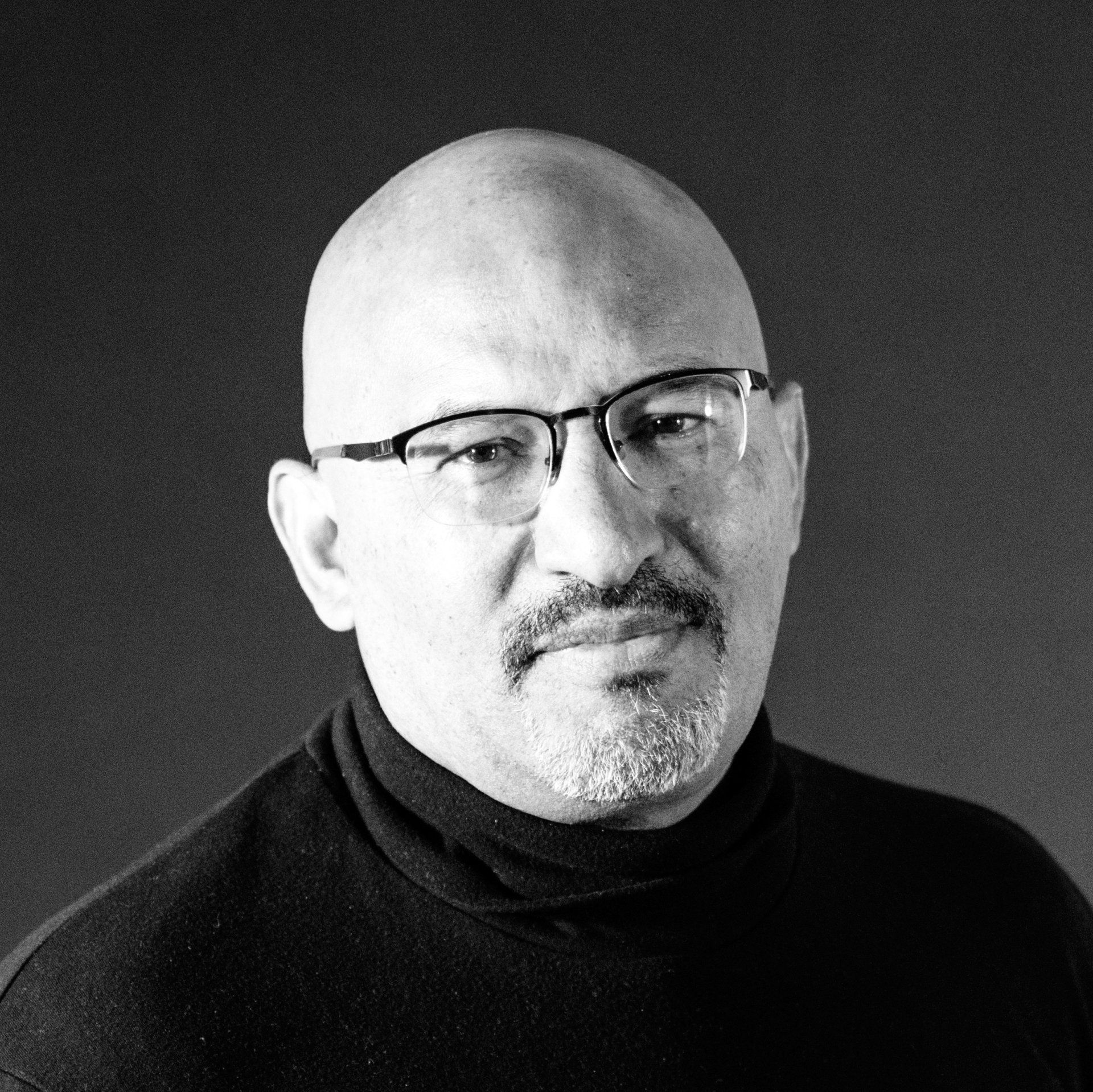 A bald man with glasses and a beard is wearing a black turtleneck.