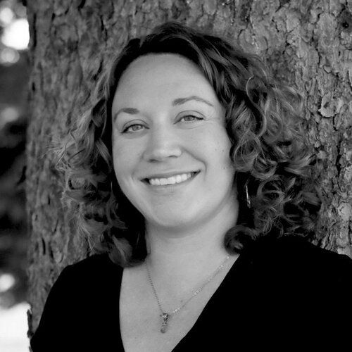 A woman is smiling in a black and white photo while leaning against a tree.
