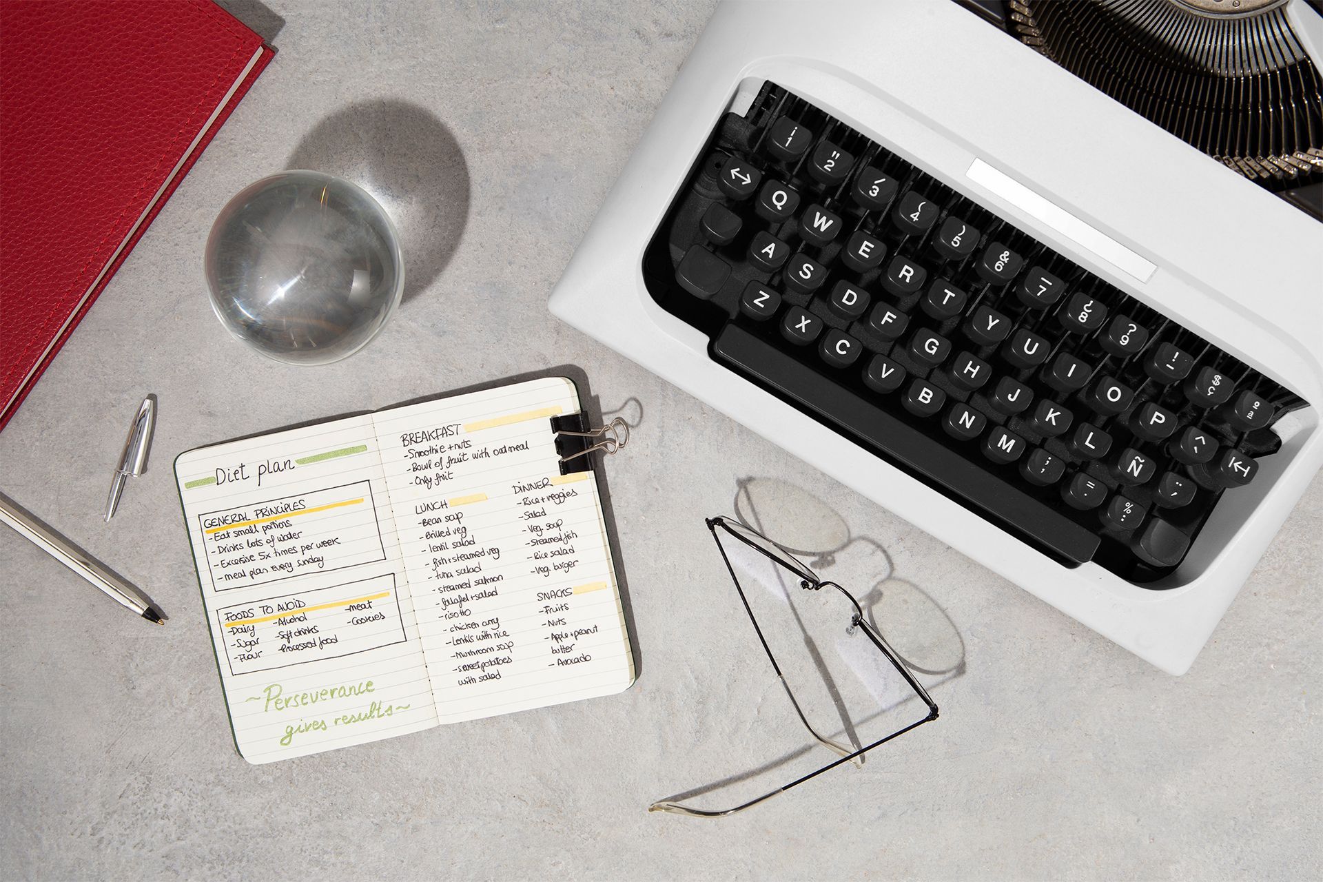 Desk with a typewriter, notebook, pen, red journal, and clear glass ball.