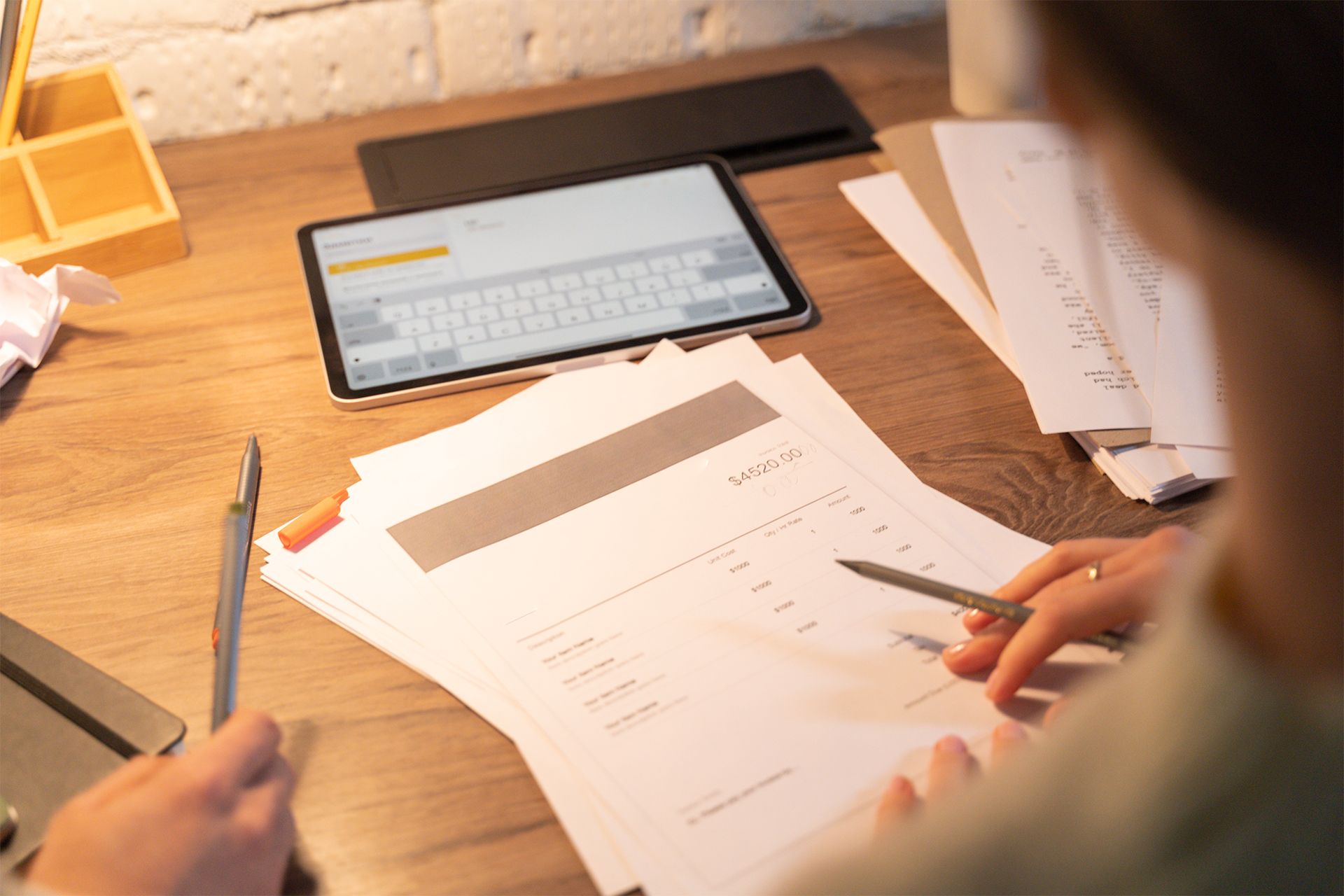 Person working at a desk with papers, a tablet showing a keyboard, and pens.